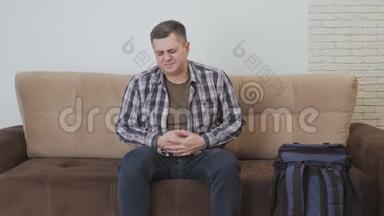 一个中年男人坐在沙发上，抱着他的胃，痛苦地<strong>扭动</strong>着。 沙发旁边是一个大型旅游背包。
